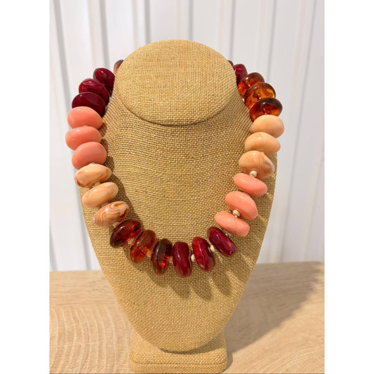A chunky beaded necklace displayed on a tan fabric jewelry bust. The necklace features alternating strands of large polished coral-pink beads, deep red marbled beads, and translucent amber-orange beads, separated by tiny white spacer beads.