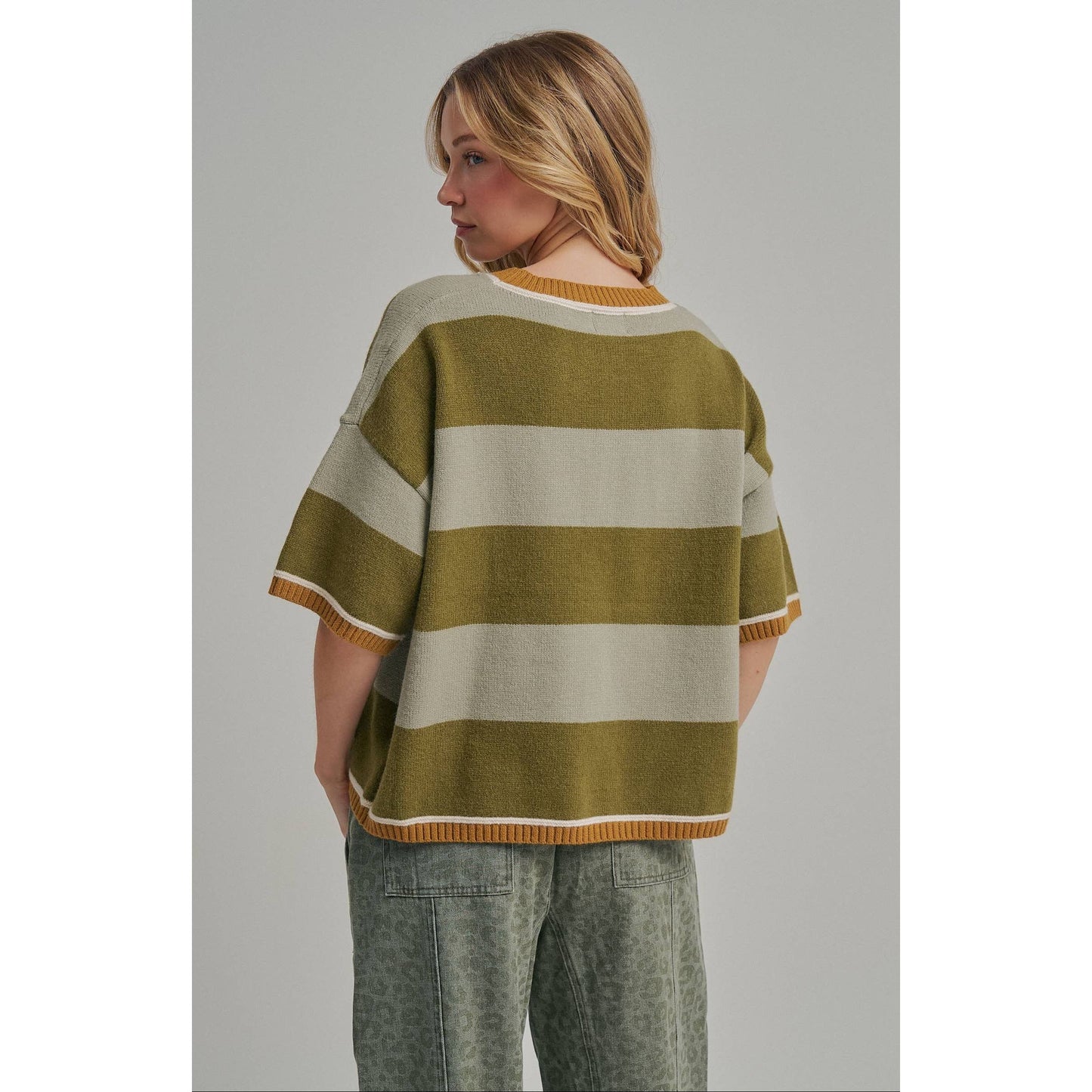 A woman shown from the back wearing a short-sleeve knit sweater with wide horizontal stripes in olive green and cream, featuring a ribbed crew neck, sleeve cuffs, and hem. She is also wearing patterned green pants against a solid gray studio background.