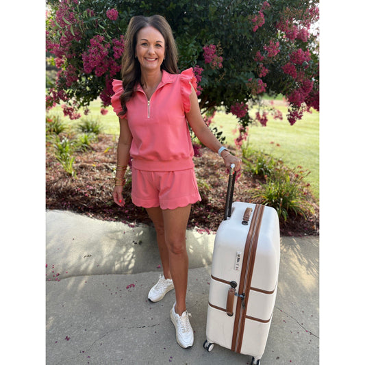 Woman in pink outfit with a white suitcase standing outdoors near a flowering bush.