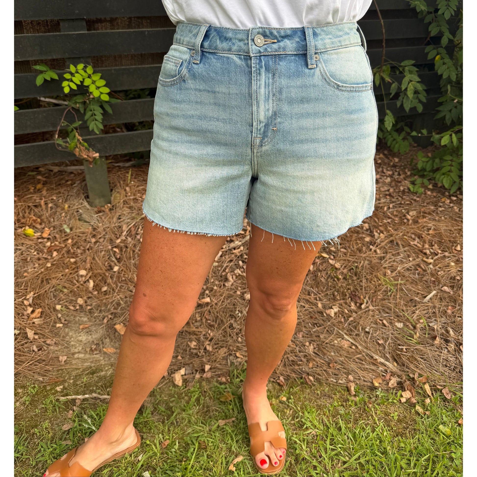 A person wearing light-wash blue denim shorts with a frayed hem, paired with a white top and tan slide sandals, standing outdoors on grass in front of a dark wooden fence.