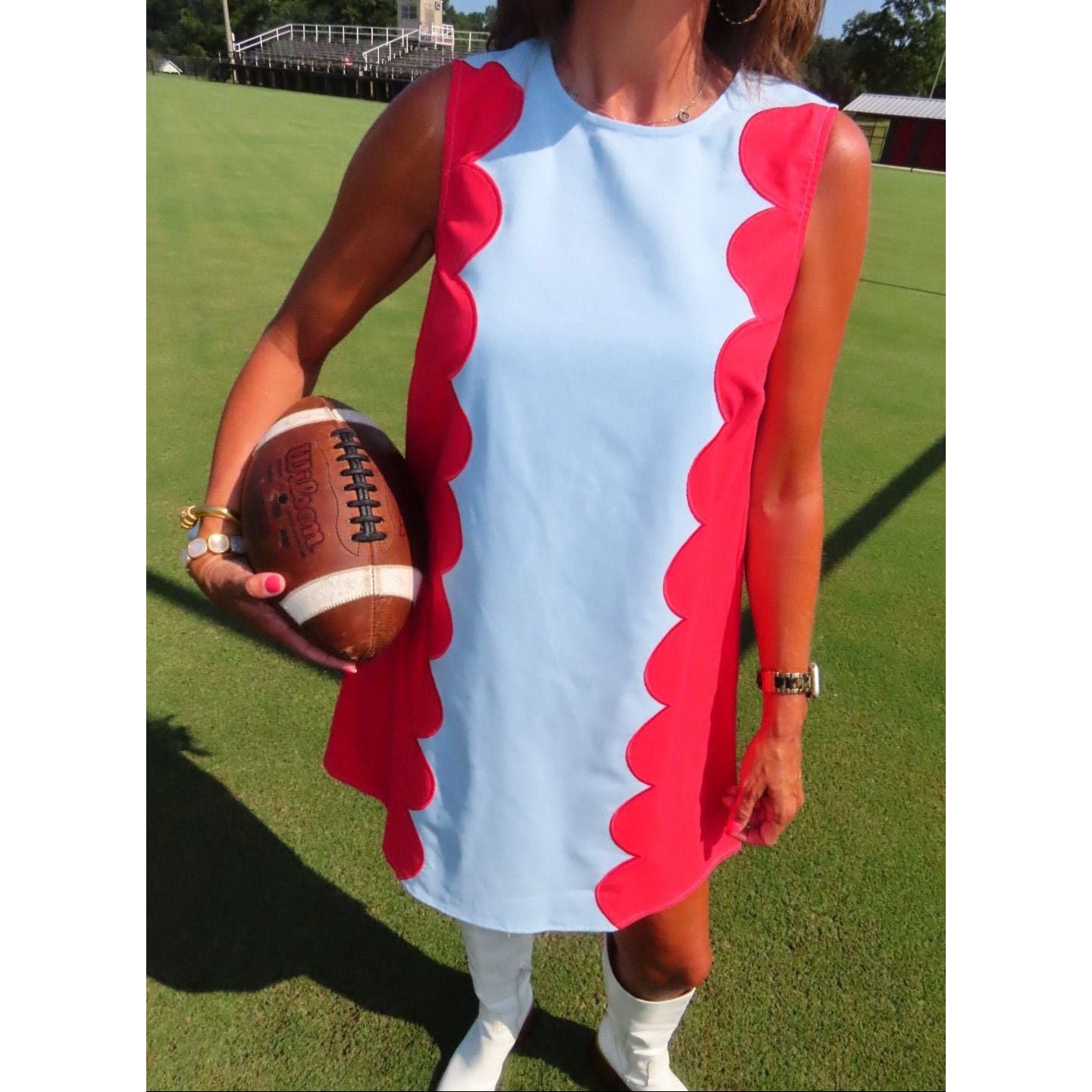 A woman on a grassy field wearing a sleeveless light blue dress with bold red scalloped side panels, holding a brown football.