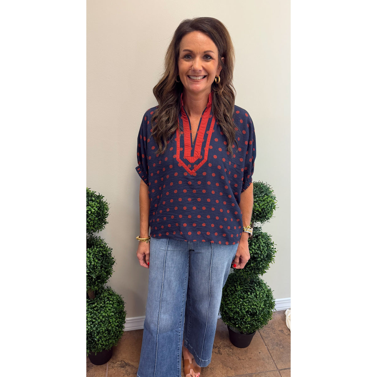 A woman stands indoors wearing a loose navy top with red polka dots and a red embroidered V-neckline, paired with light blue wide-leg jeans. She is barefoot on a tiled floor, with two green topiary trees on either side against a beige wall.