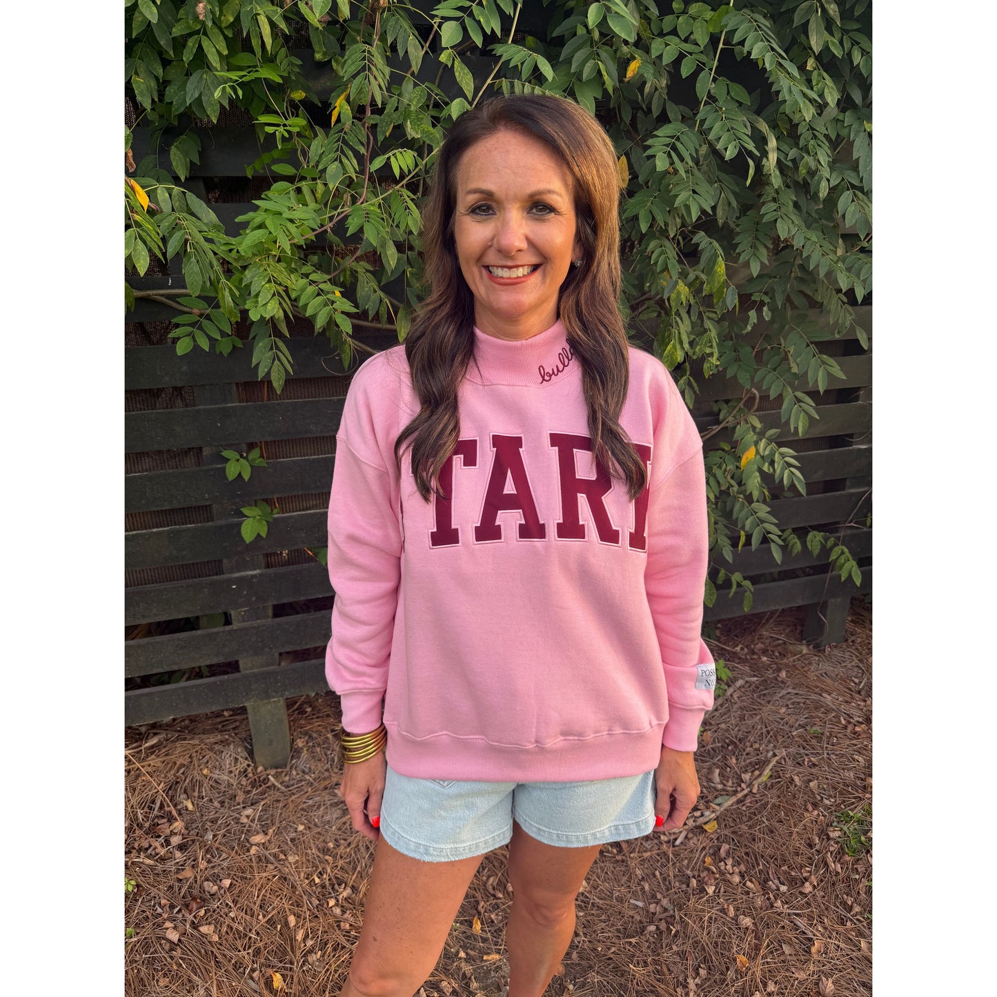 A woman wearing a pink sweatshirt with 'STARK' printed in maroon across the chest, paired with light blue denim shorts, standing outdoors in front of a wooden fence and green foliage.