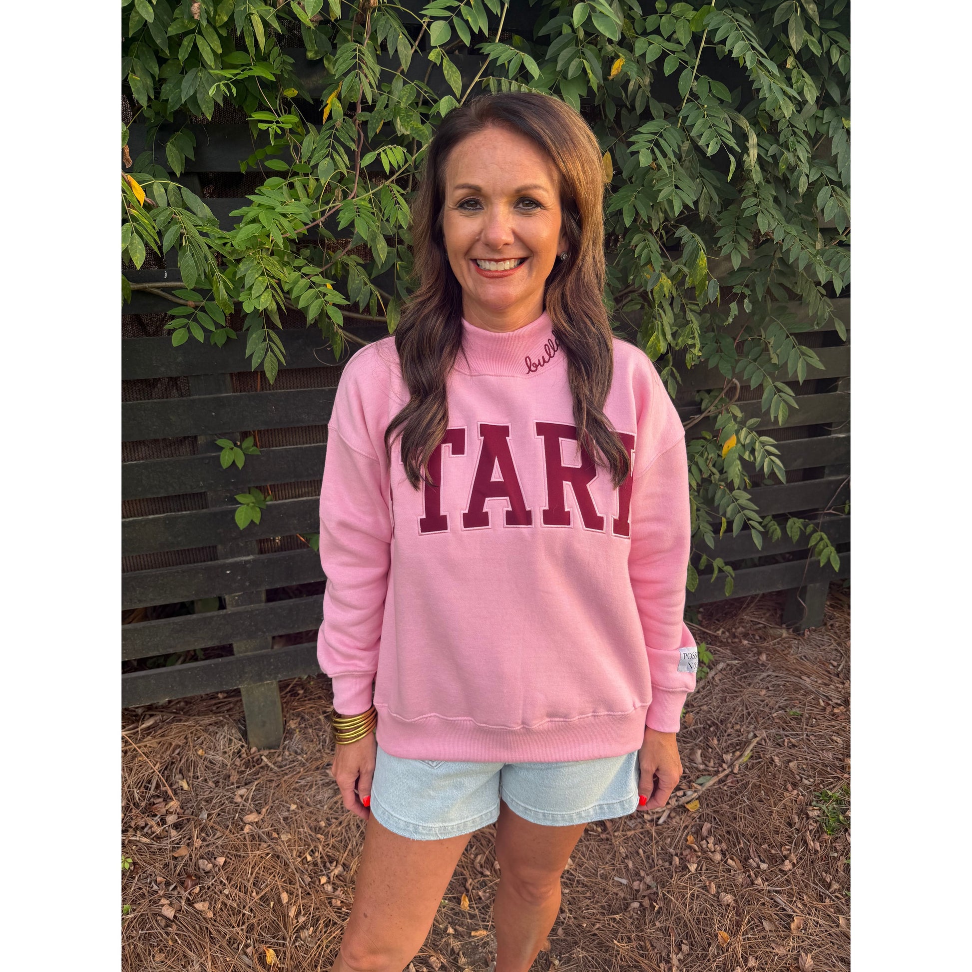 A woman wearing a pink sweatshirt with 'STARK' printed in maroon across the chest, paired with light blue denim shorts, standing outdoors in front of a wooden fence and green foliage.