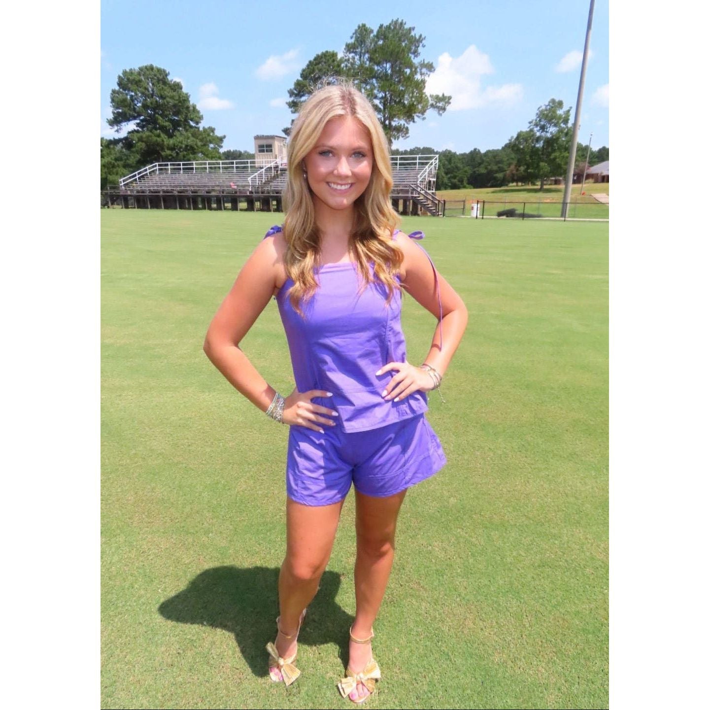 A woman stands on a grassy field wearing a matching purple sleeveless top and shorts set, with trees and bleachers in the background.
