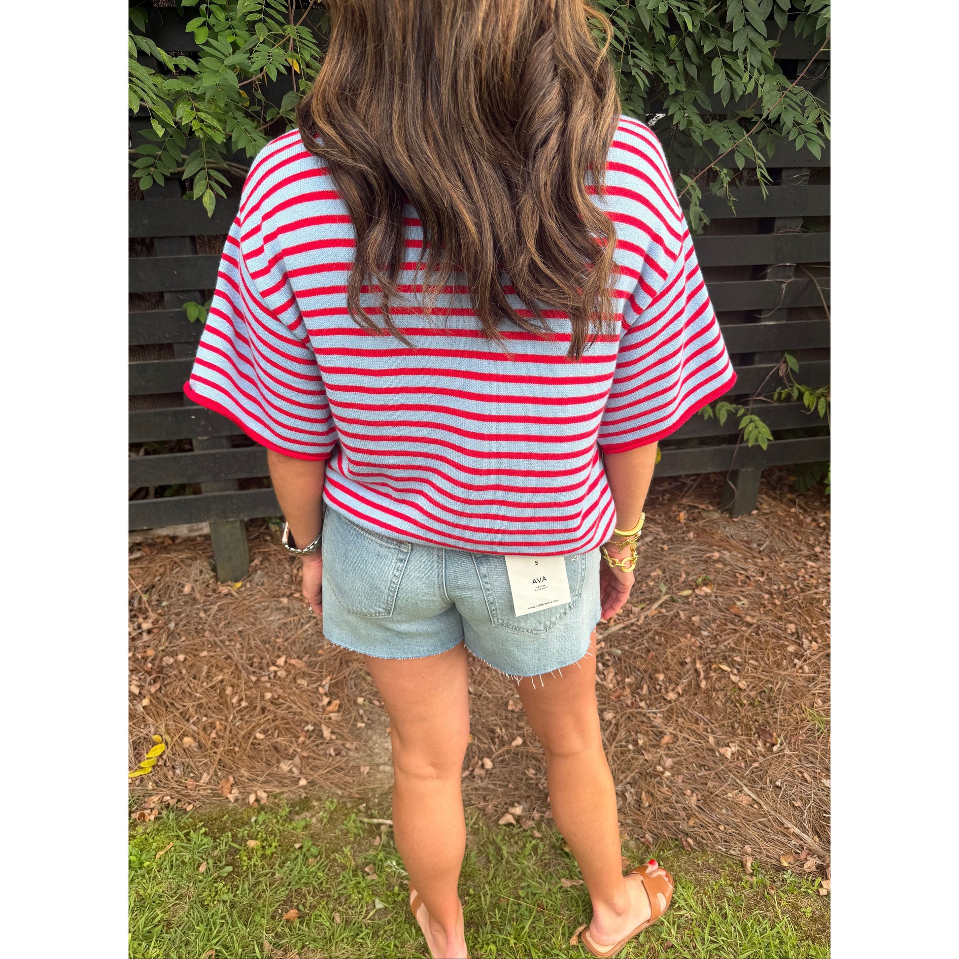 A woman viewed from behind wearing a red and white striped short-sleeve t-shirt, denim shorts, and sandals, standing outdoors in front of a dark wooden fence.