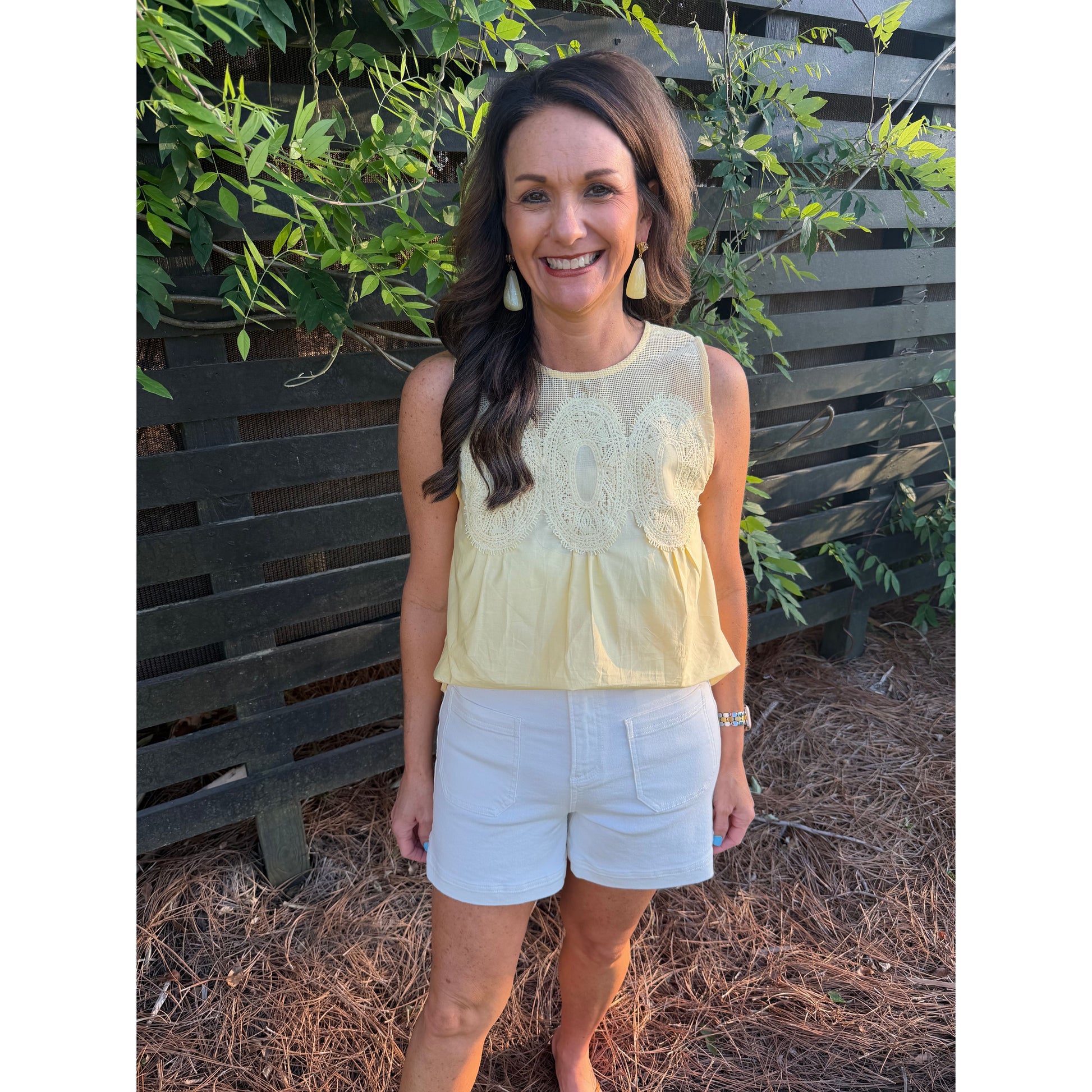 Woman wearing a yellow top and white shorts standing in front of a wooden fence with greenery.