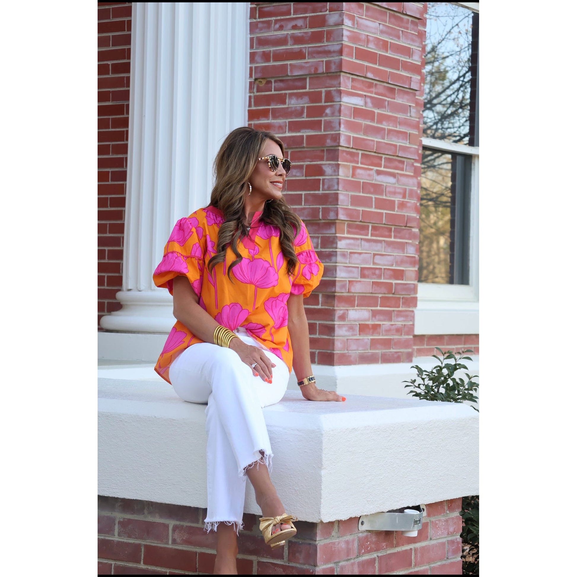 Woman in a colorful shirt and white pants sitting on a white bench against a brick wall.