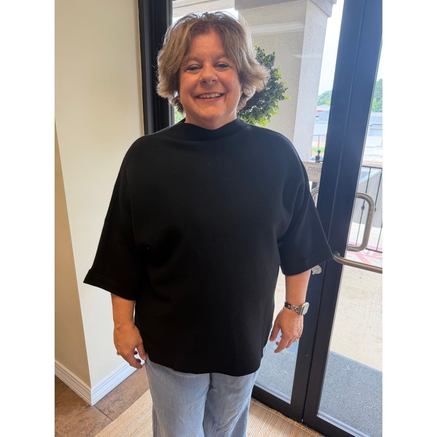 A smiling woman with short gray hair wearing a black mock-neck short-sleeve top and light blue jeans, standing indoors near a glass door.
