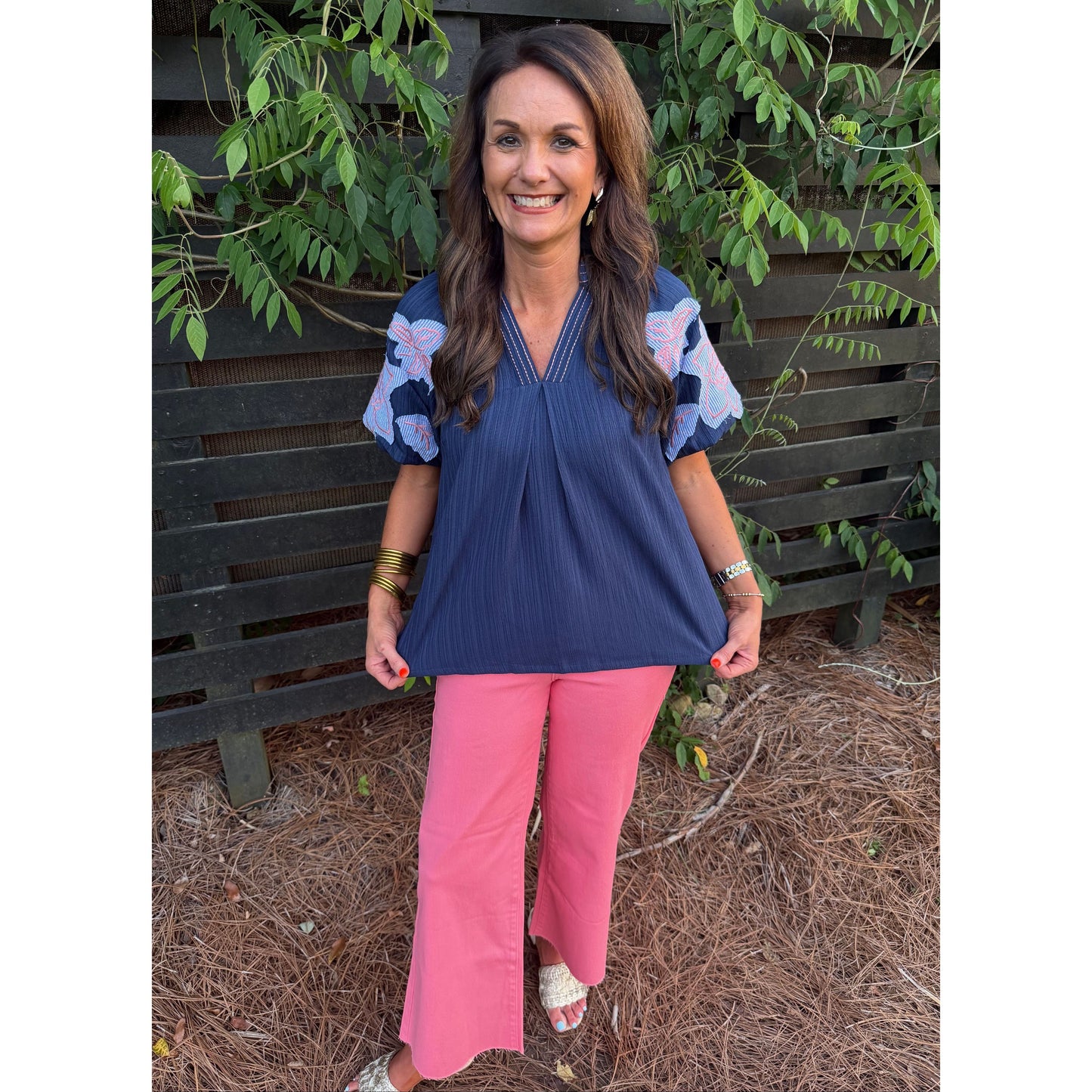Woman wearing a blue top and pink pants standing outdoors with greenery in the background