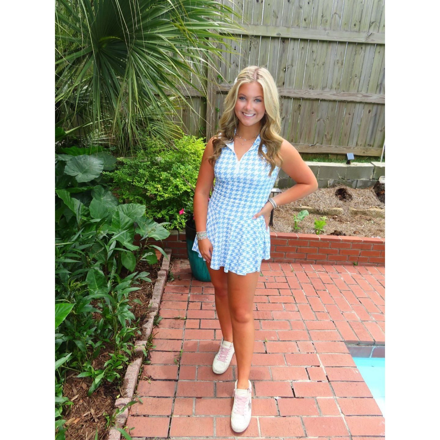 Woman in a checkered dress standing on a brick patio with greenery and a wooden fence in the background.