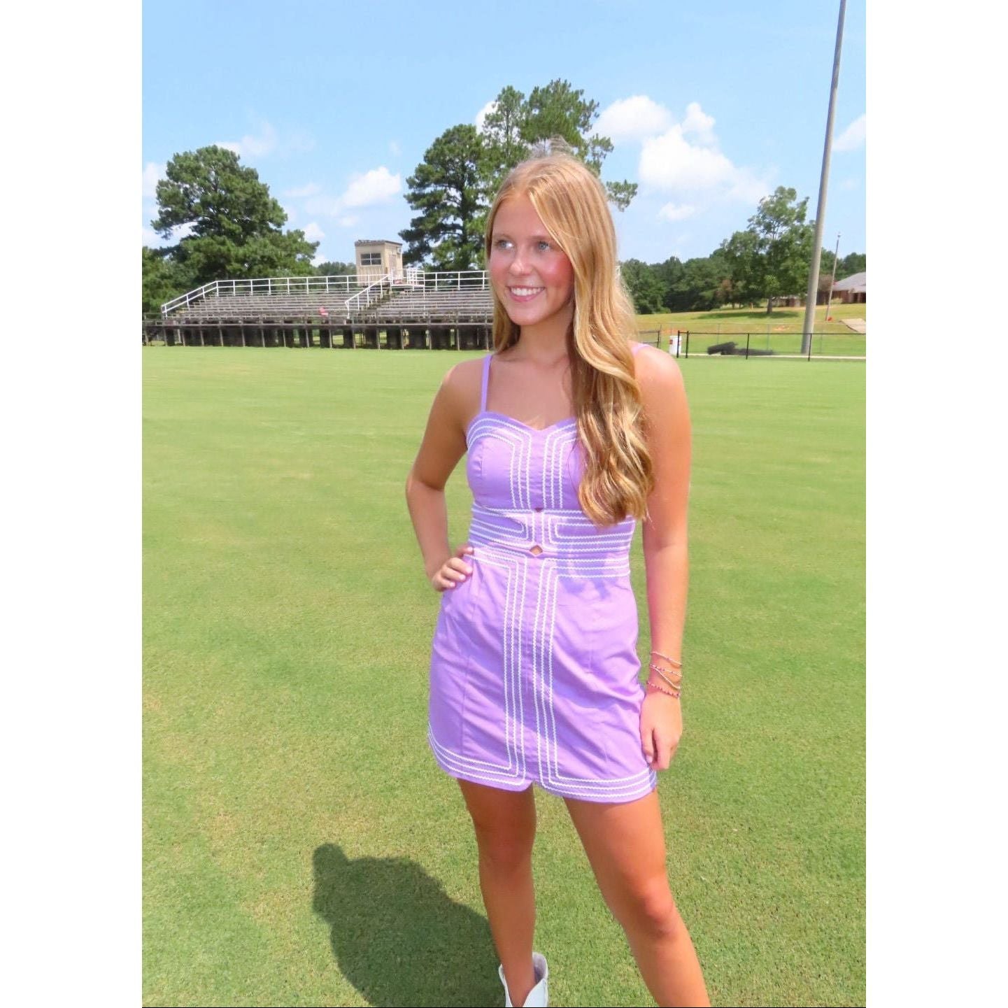 A young woman with blonde hair stands on a green grass field wearing a lavender and white striped mini dress. In the background, there is a large wooden bleacher structure and trees under a blue sky with light clouds.