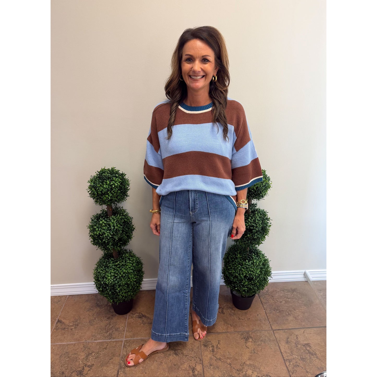 A woman stands indoors wearing a brown and light blue striped knit sweater with wide sleeves, paired with wide-leg blue jeans and tan flat sandals. She is positioned between two small green topiary trees against a beige wall.