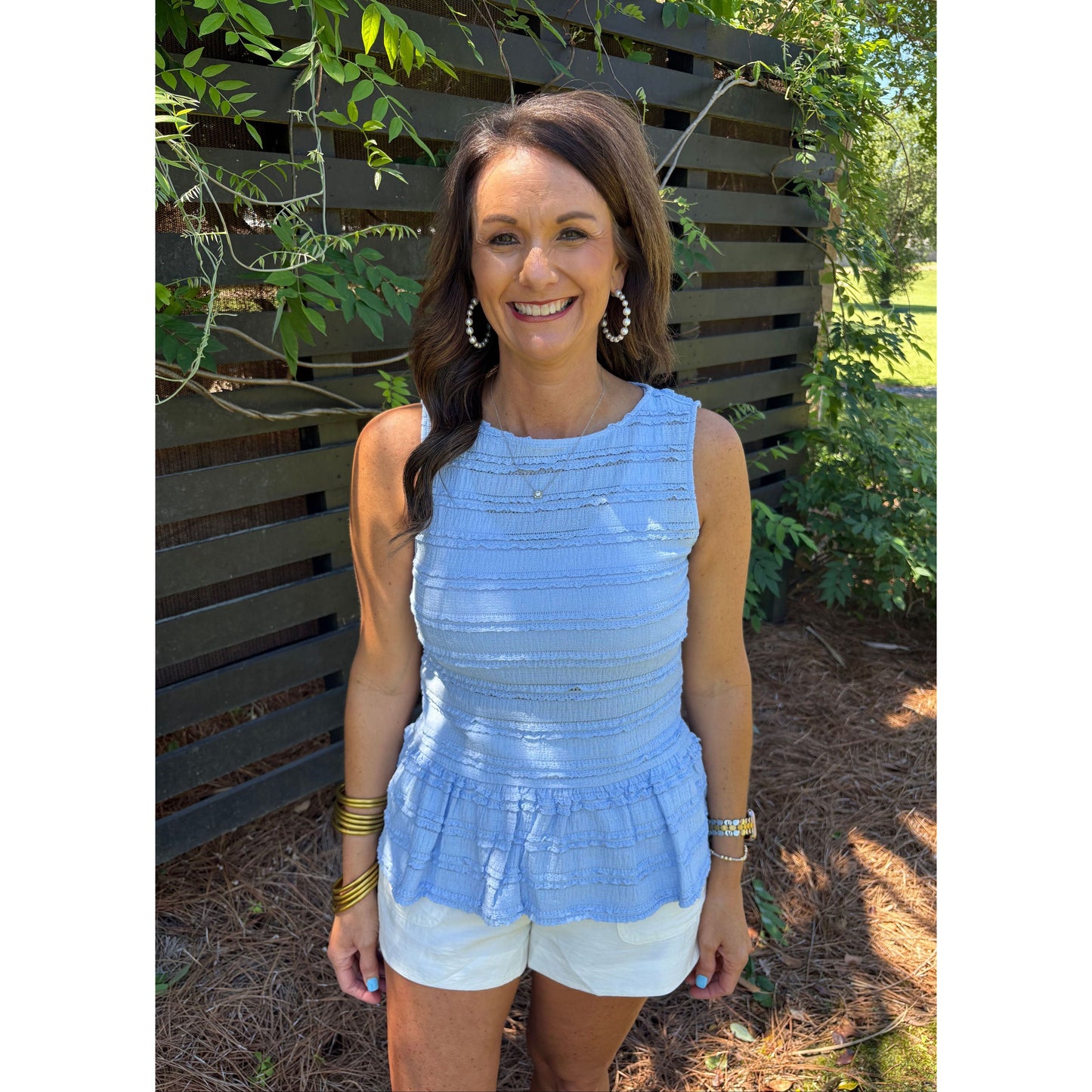 Woman wearing a light blue sleeveless top and white shorts standing outdoors with greenery in the background.