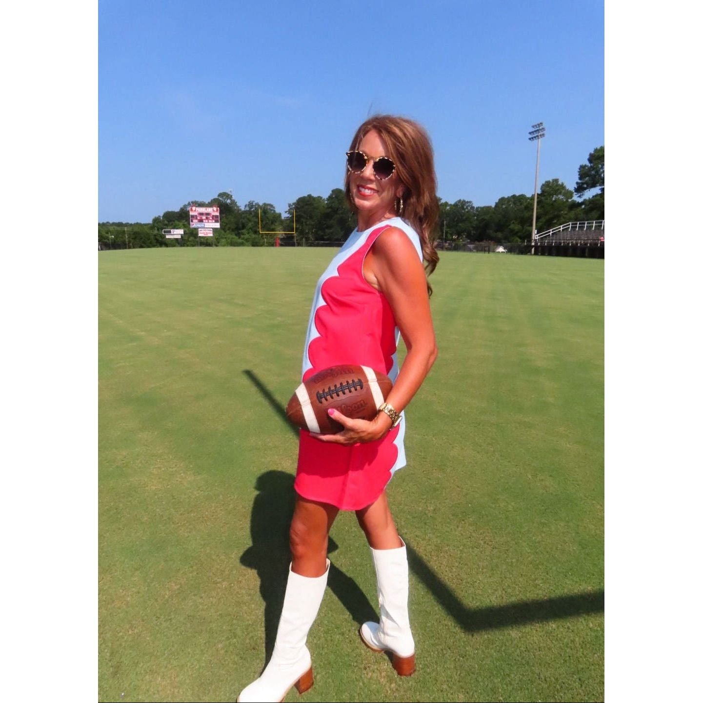 A woman stands on a grassy field holding a football, wearing a sleeveless pink and white retro-style dress with white knee-high boots and sunglasses.
