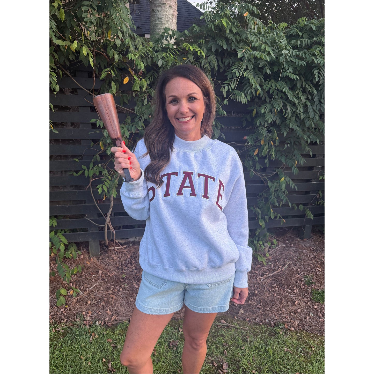 A woman wearing a light blue sweatshirt with 'STATE' printed in maroon across the chest, paired with light blue denim shorts, standing outdoors in front of a dark fence and greenery while holding a wooden mallet.