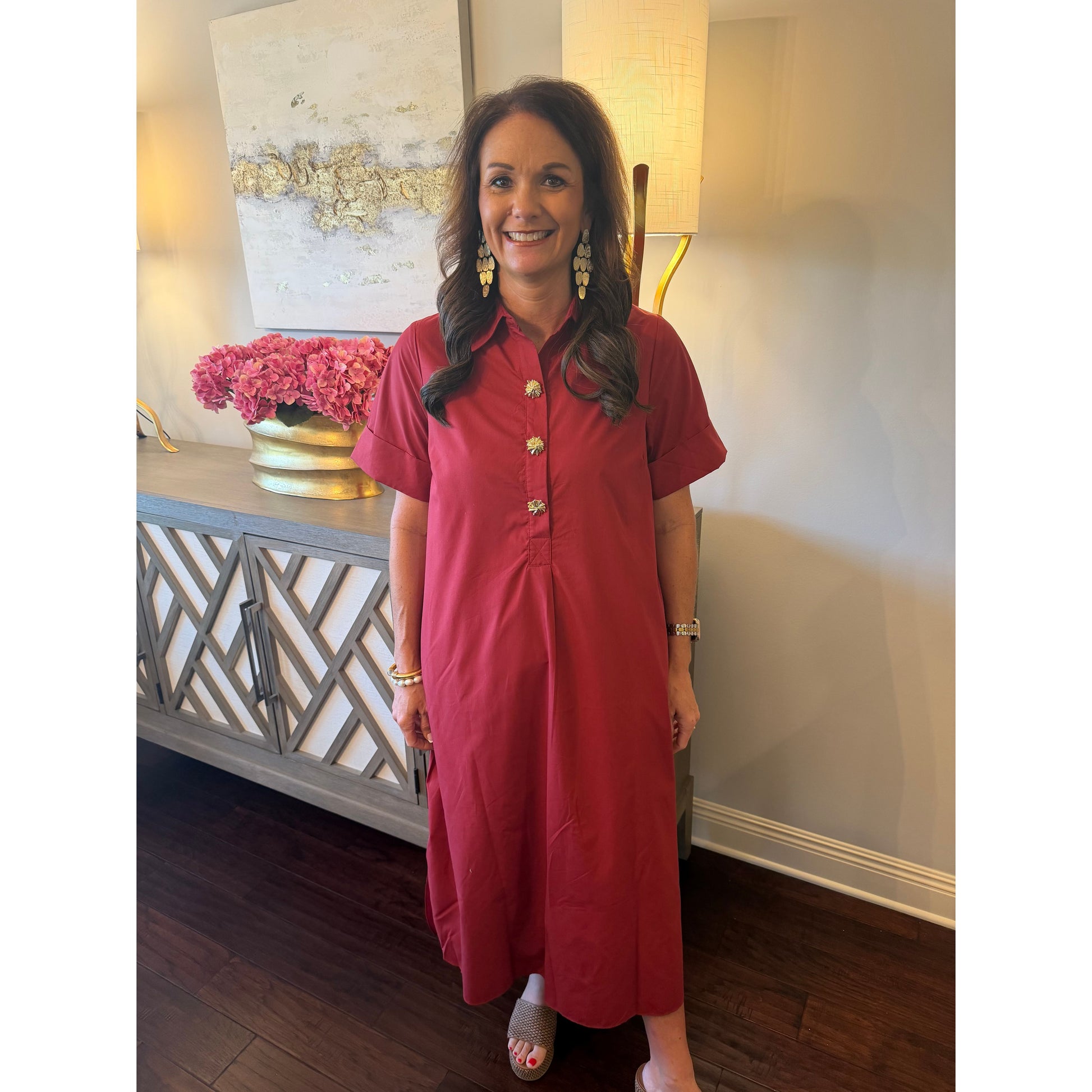 A woman wearing a short-sleeve red button-down dress with gold buttons, standing in a home interior setting with a console table and lamp in the background.