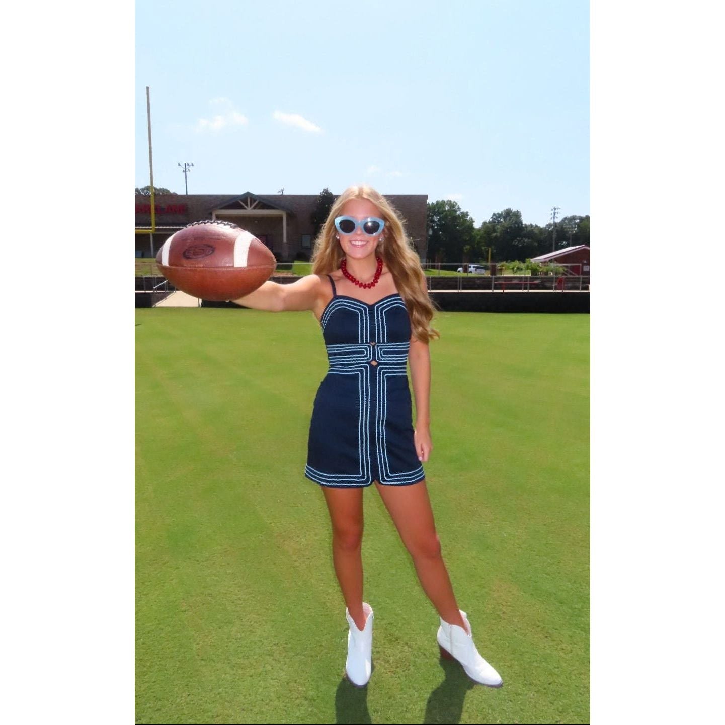 A woman stands on a green grass field outdoors, wearing a navy blue sleeveless mini dress with white contrast piping, white ankle boots, and white sunglasses while holding a brown football.