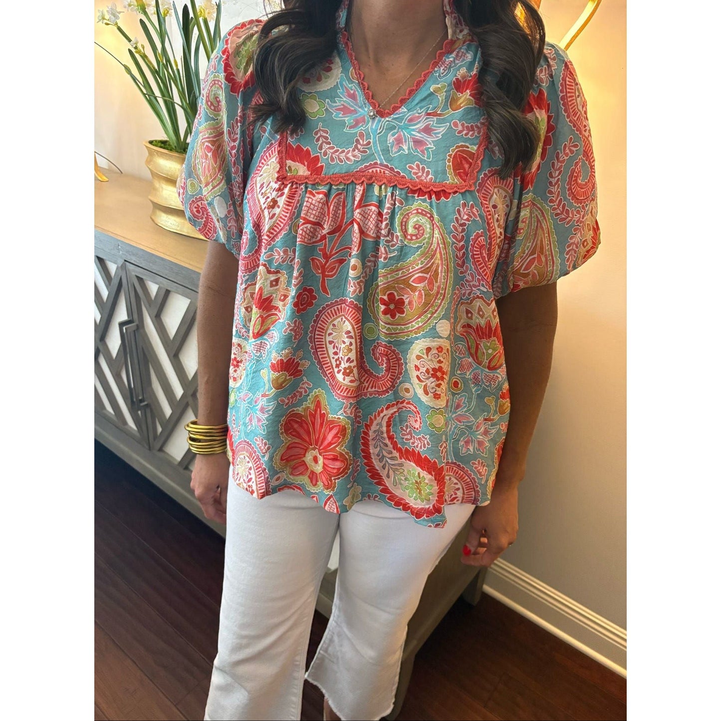 Person wearing a colorful paisley blouse with white pants indoors.