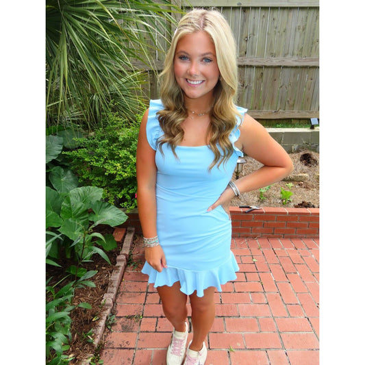 Woman in a light blue dress standing outdoors on a brick path with greenery.