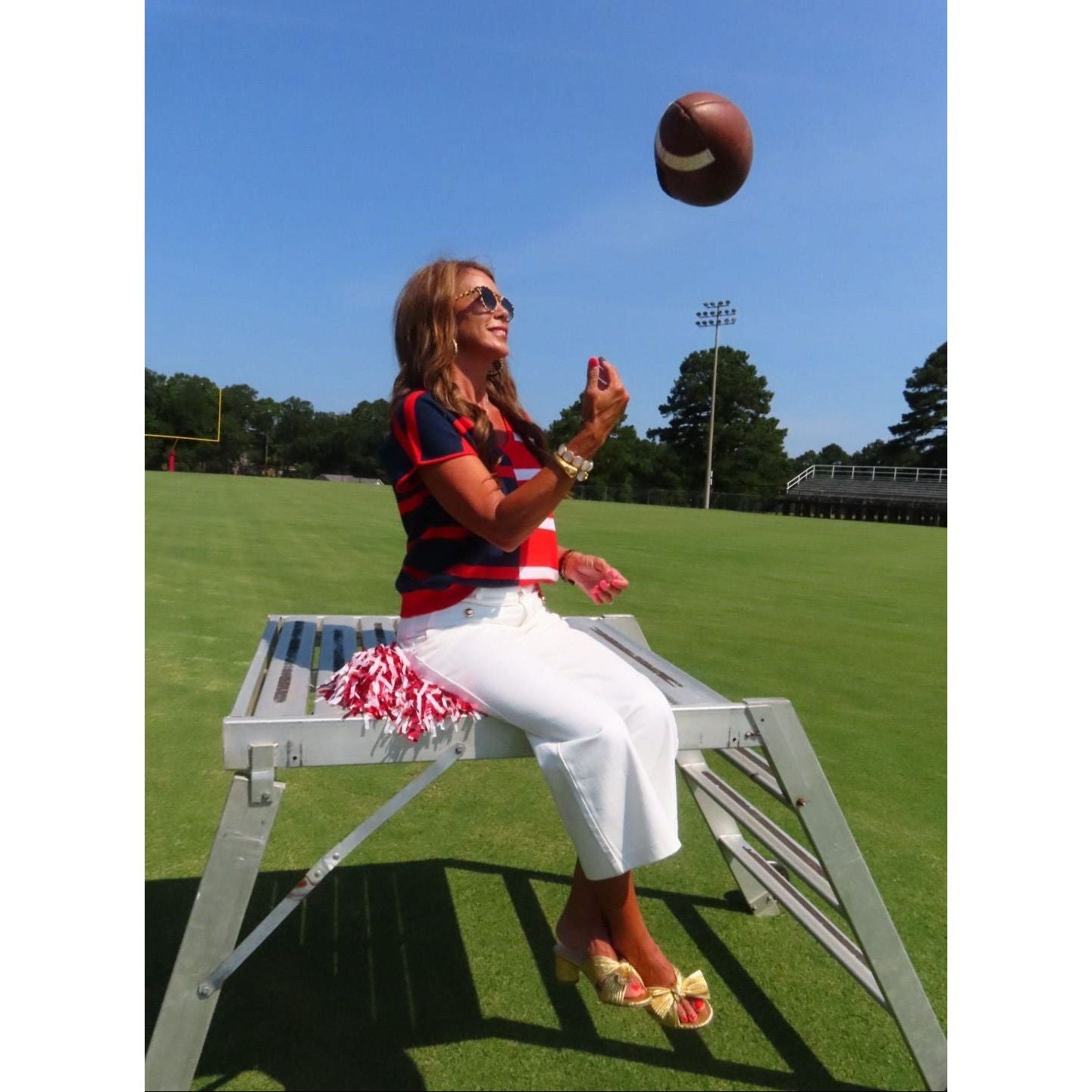 A woman sitting on a metal football field chair on a grassy field, wearing a red and navy striped shirt, white pants, and yellow sandals, with a football floating in the air against a blue sky.