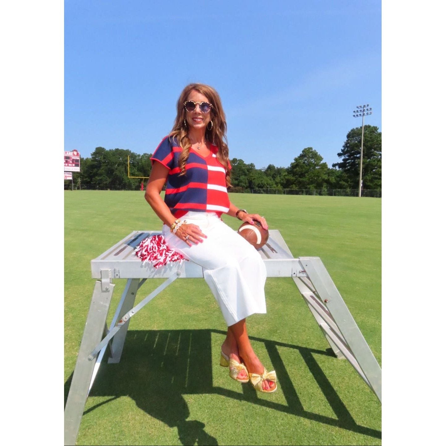 A woman wearing a red, navy, and white striped short-sleeve top, white pants, and beige sandals sits on a white metal bench on a green grass field under a blue sky.