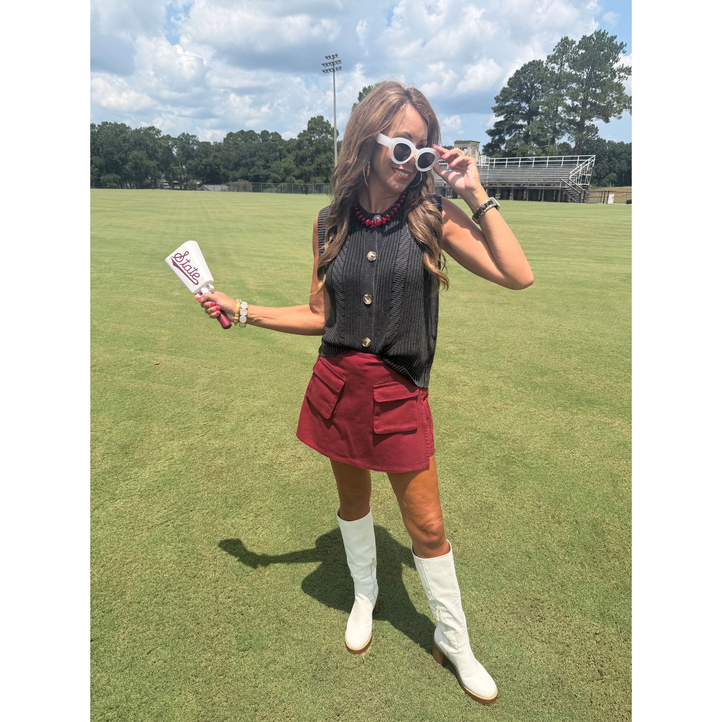 A woman stands on a grassy field wearing a black ribbed sleeveless top, a red mini skirt, white knee-high boots, and white sunglasses. She is holding a white paddle with red text that reads 'Sizzle'.
