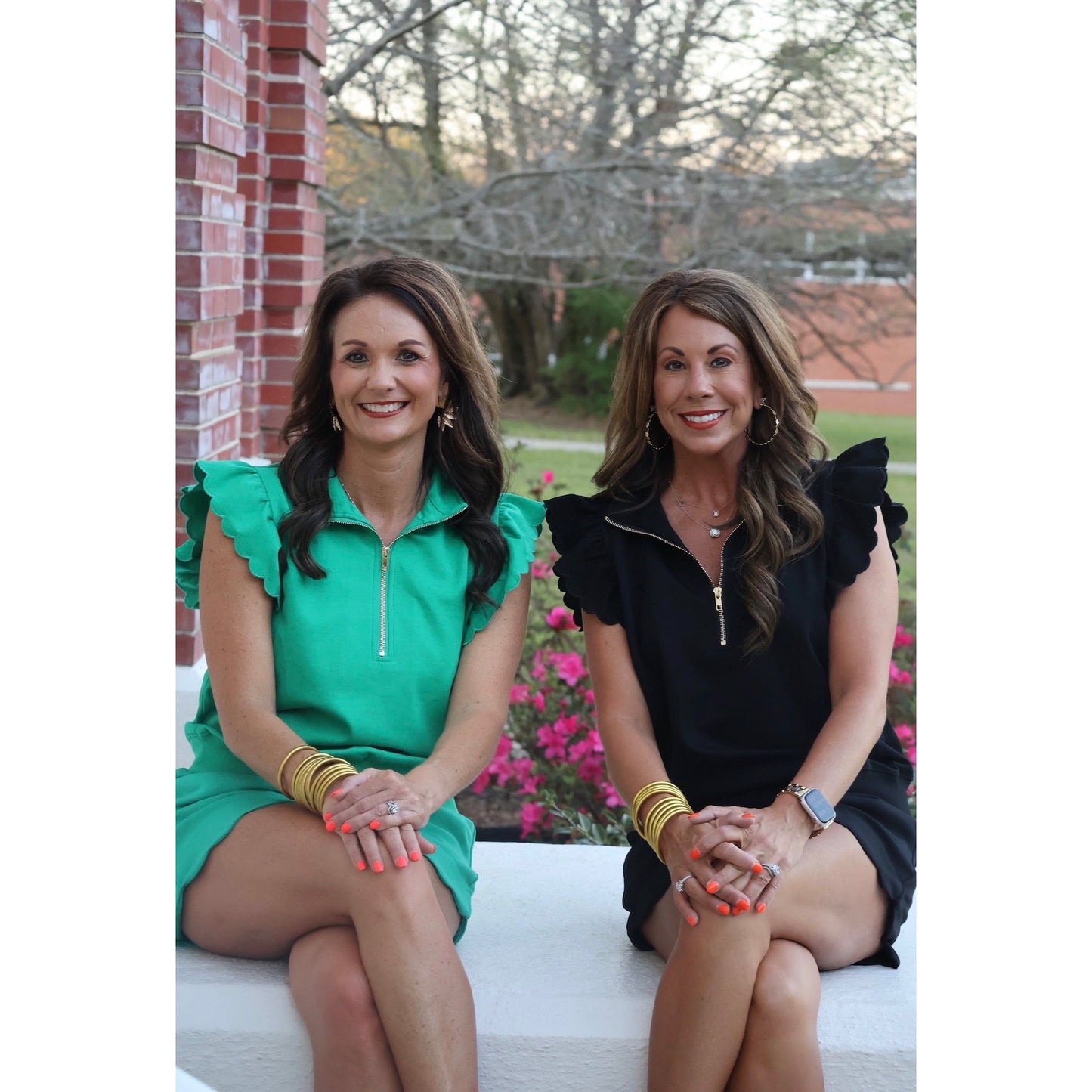 Two women sitting on a bench outdoors with flowers and a brick wall in the background.