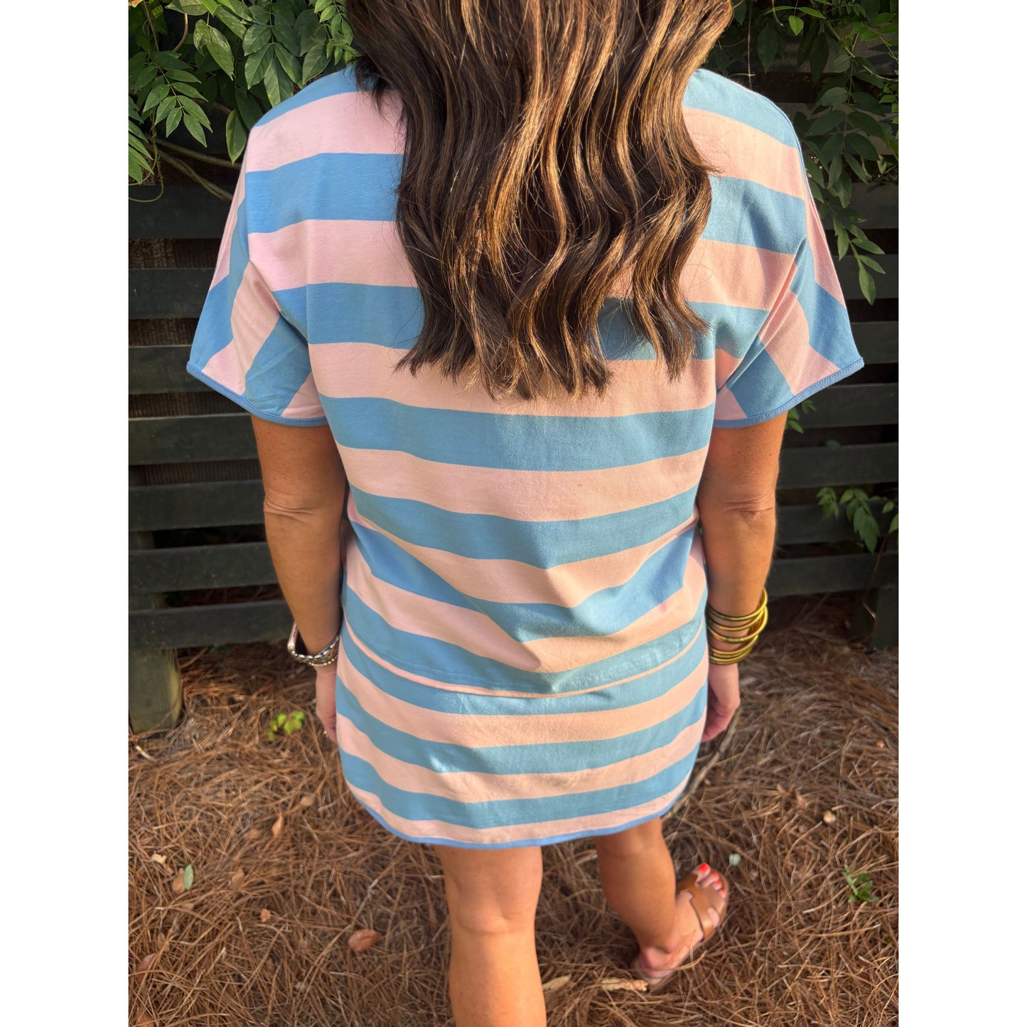 Person wearing a blue and pink striped dress standing outdoors with greenery in the background