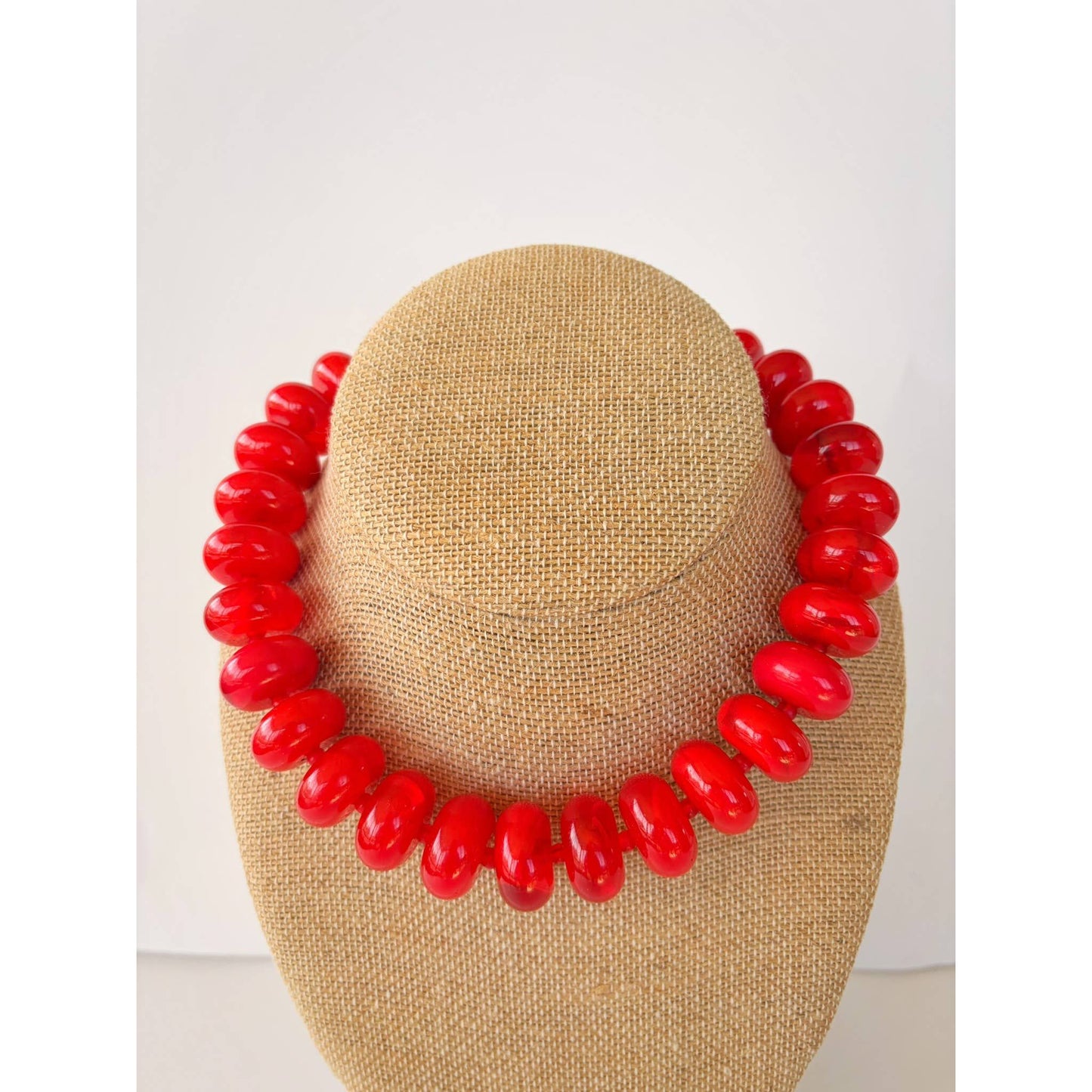 A bright red beaded necklace displayed on a tan burlap-covered jewelry bust against a plain white background.