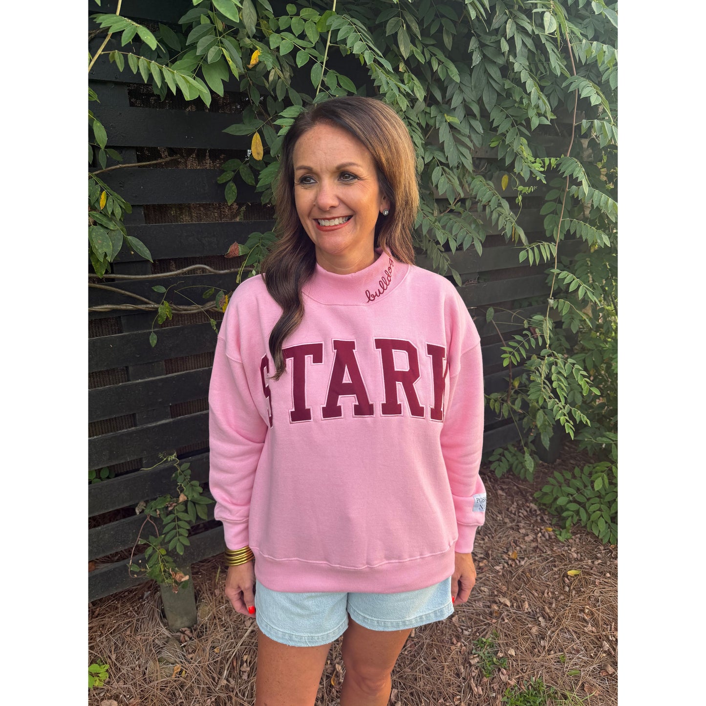 A woman wearing a pink sweatshirt with 'STARD' printed in maroon across the front, standing outdoors in front of a dark wooden fence and green plants.
