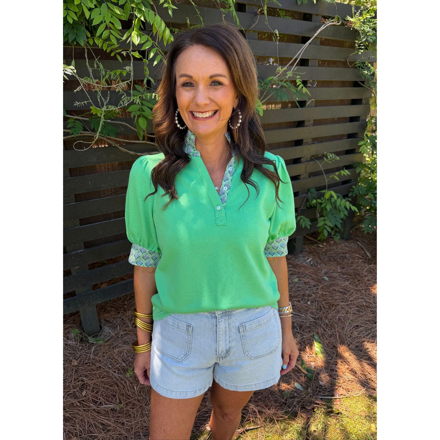 Woman in a green shirt and denim shorts standing outdoors with plants and a wooden fence in the background.