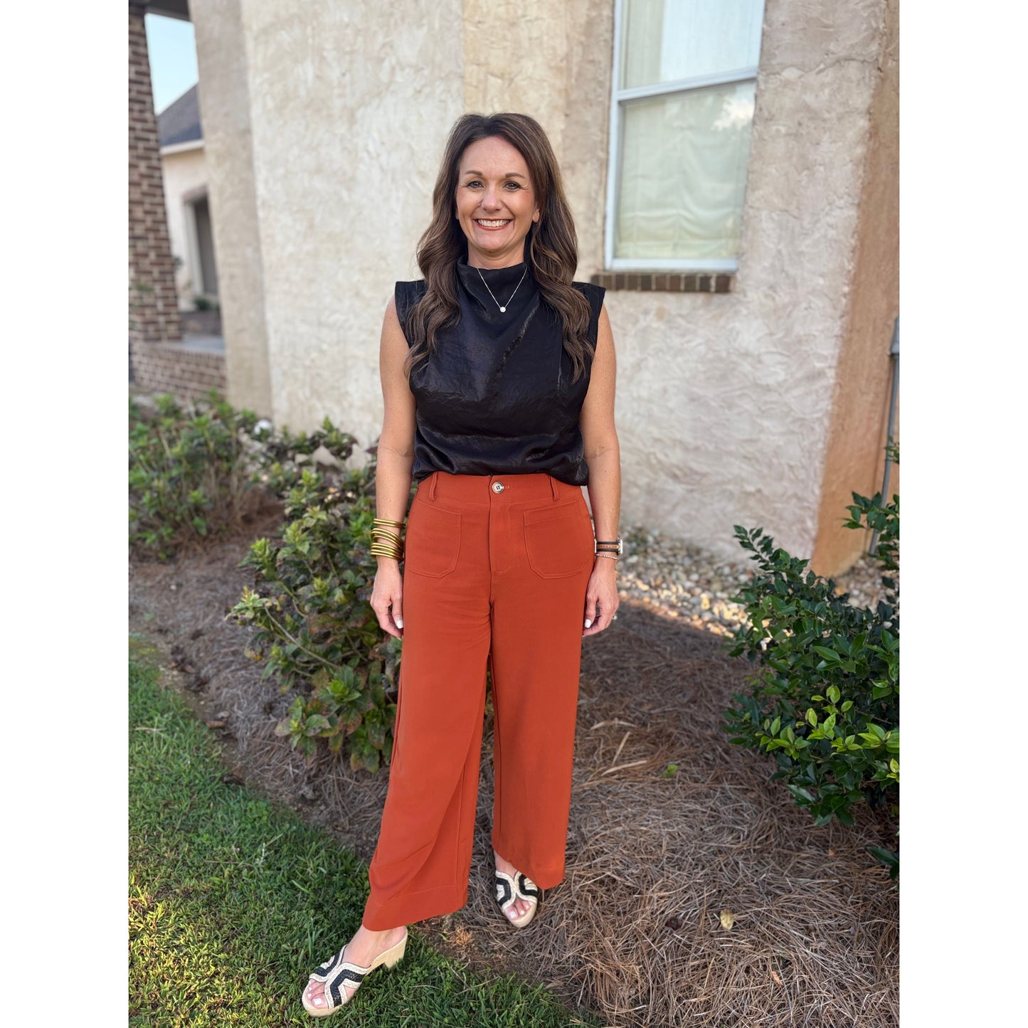 Woman wearing a black sleeveless top and rust-colored pants standing outside a house.