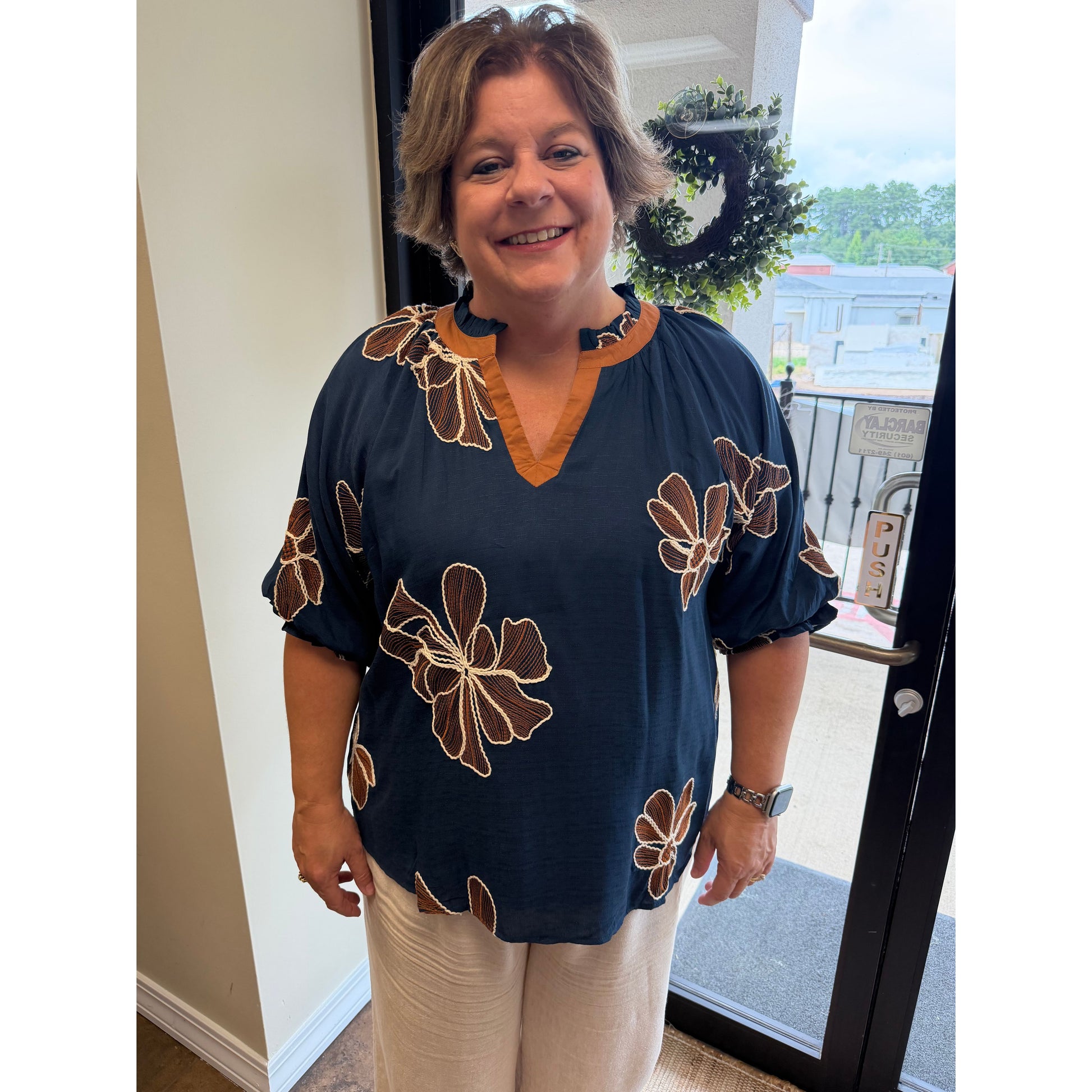 A woman wearing a navy blue short-sleeve blouse with brown and beige floral patterns and a tan V-neck trim, paired with beige pants, standing in front of a glass door.