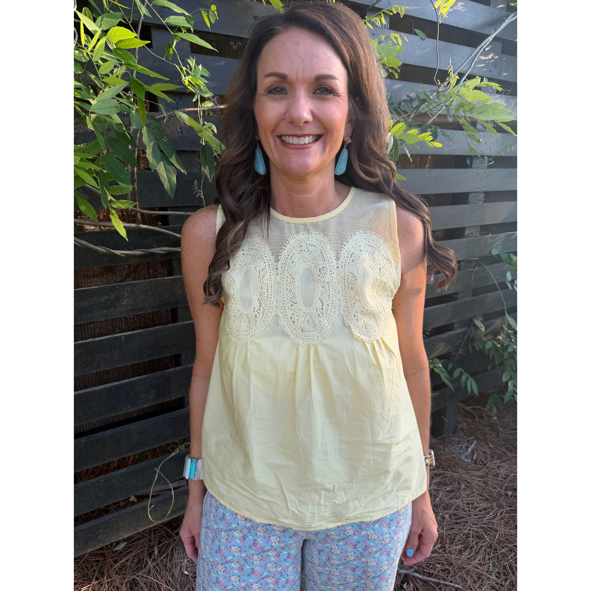 Woman wearing a light yellow sleeveless top with lace details, standing outdoors against a wooden fence.