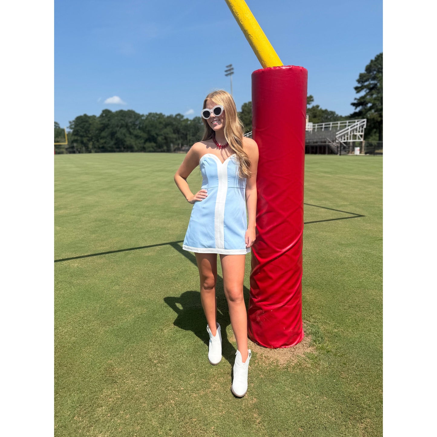 A woman wearing a light blue and white striped mini dress and white sneakers stands on a grassy field next to a red and yellow football goal post.