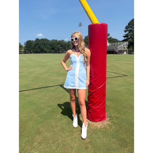 A woman wearing a light blue and white striped mini dress and white sneakers stands on a grassy field next to a red and yellow football goal post.