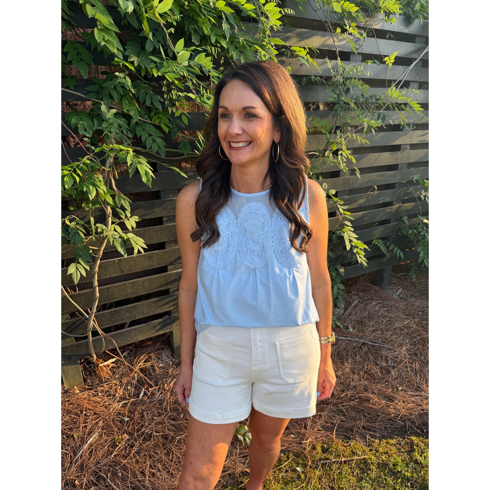 Woman in a light blue top and white shorts standing outdoors with greenery in the background