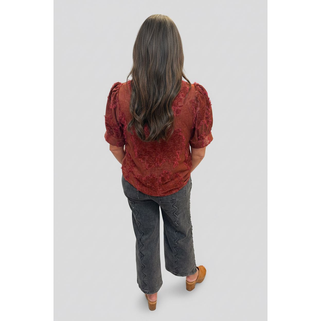 A woman viewed from behind wearing a rust-red textured short-sleeve blouse and dark gray wide-leg jeans, standing on a tiled indoor floor next to a white column.