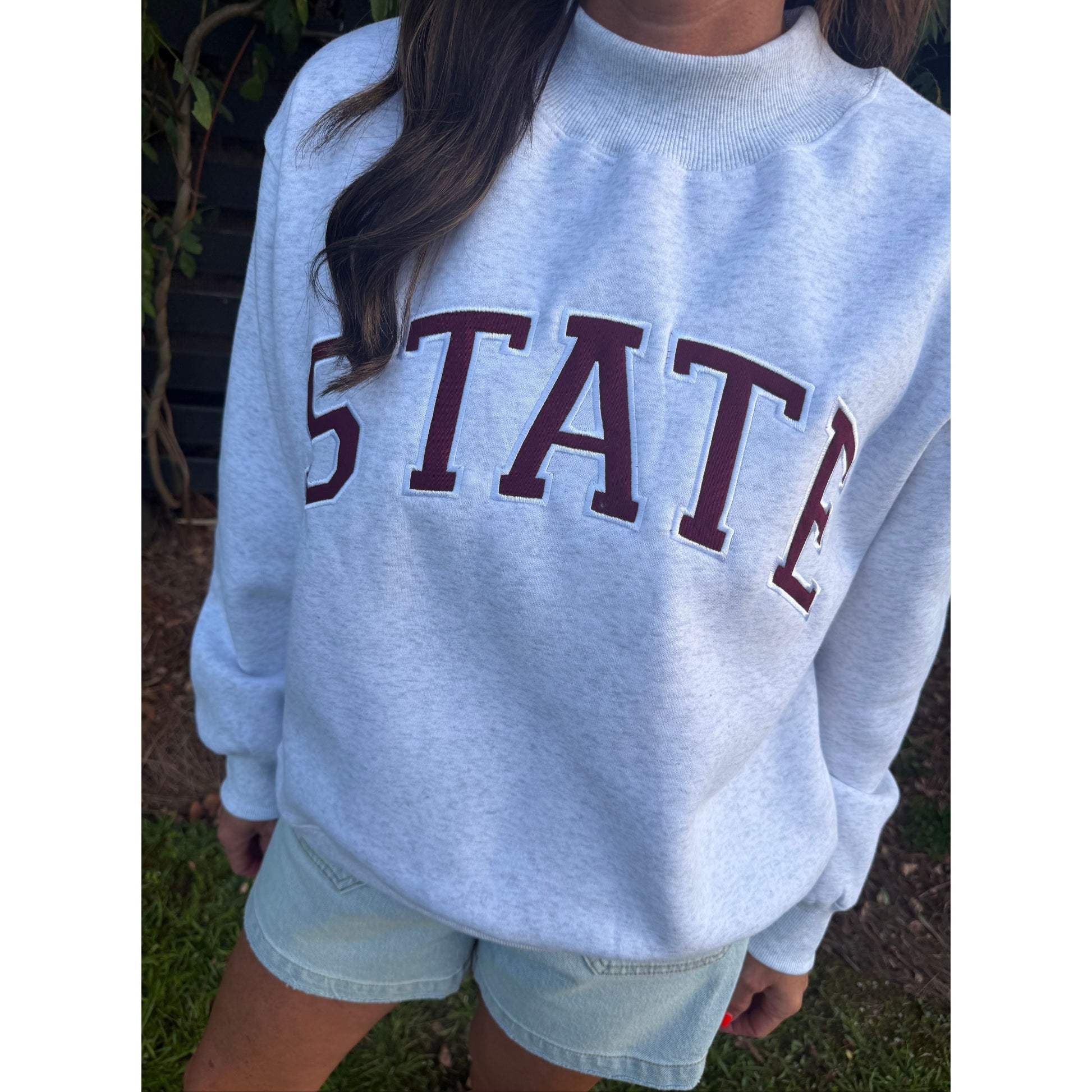 A person wearing a light heather gray sweatshirt with 'STATE' printed across the chest in dark maroon varsity letters, paired with light denim shorts, photographed outdoors on grass.