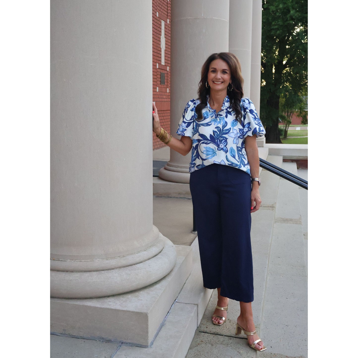 Woman in a blue floral blouse and dark pants standing outdoors near architectural columns.
