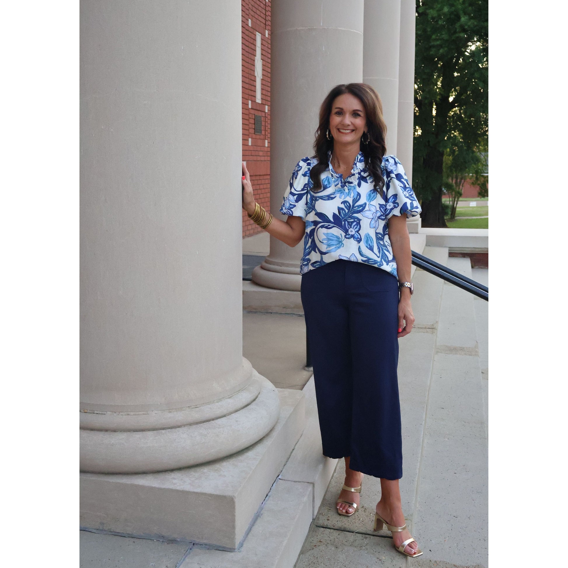 Woman in a blue floral blouse and dark pants standing outdoors near architectural columns.