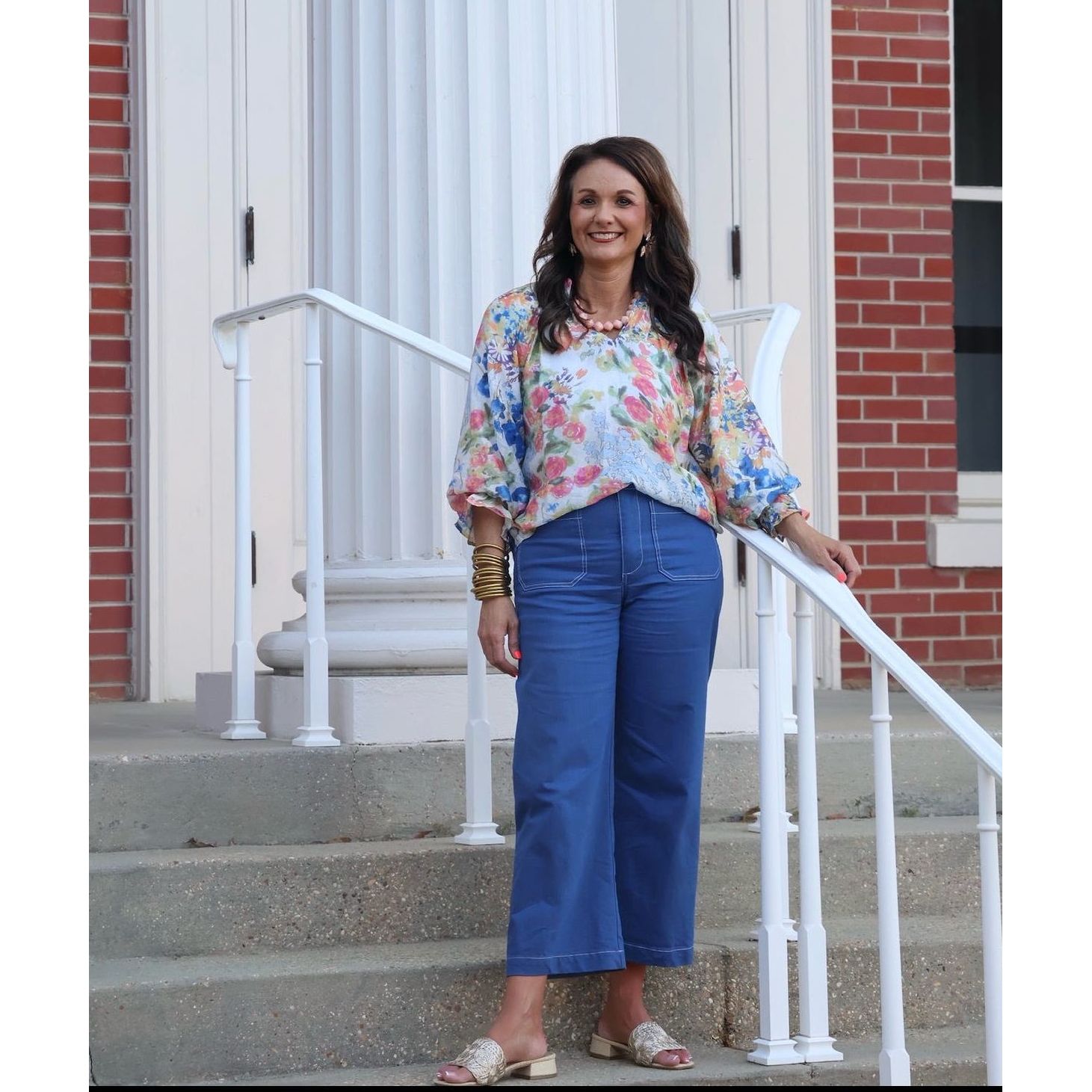 Woman standing on a set of stairs in front of a brick building wearing a floral blouse and blue pants.