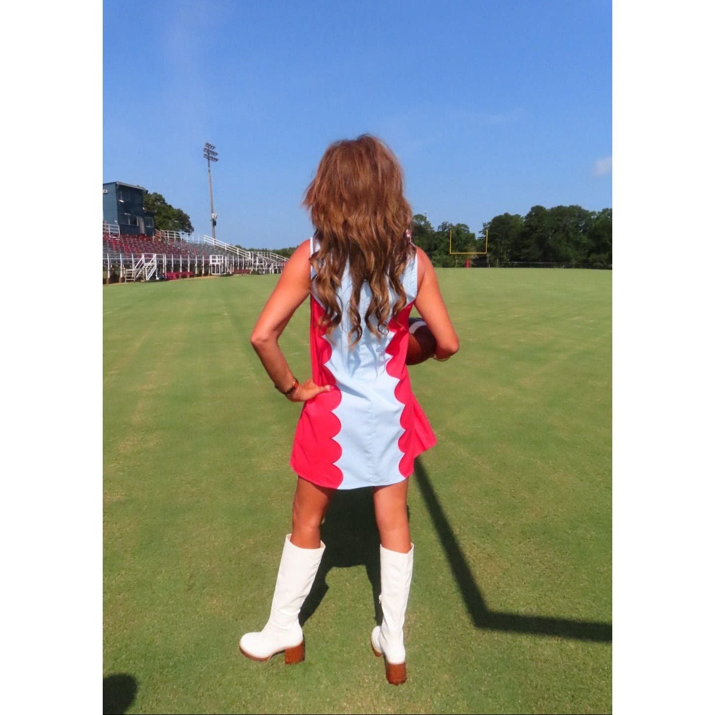 A woman stands on a green sports field wearing a light blue sleeveless dress with bright pink ruffled side panels and white knee-high boots, photographed from behind.