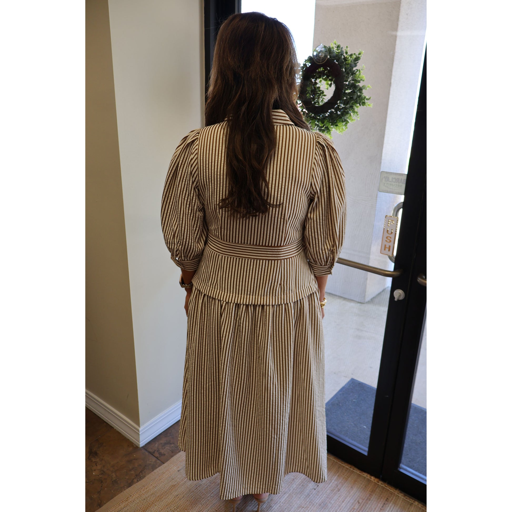A woman viewed from behind wearing a beige and brown vertically striped midi dress with puff sleeves and a tie waist, standing in an indoor doorway.