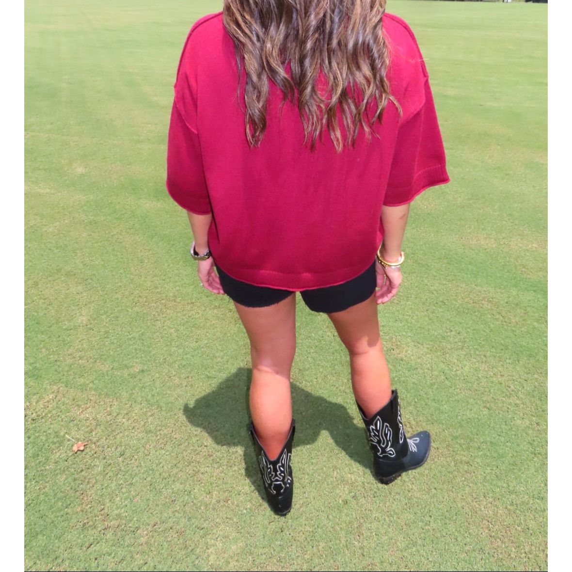 A woman viewed from behind wearing an oversized dark red t-shirt, black shorts, and black cowboy boots on a green grass field.