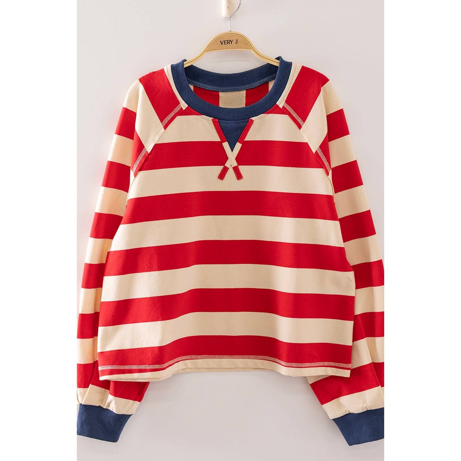 A long-sleeve raglan pullover sweatshirt with horizontal red and cream stripes, featuring a navy blue crew neck, split V-neckline, and ribbed cuffs, hanging on a wooden hanger against a light gray background.