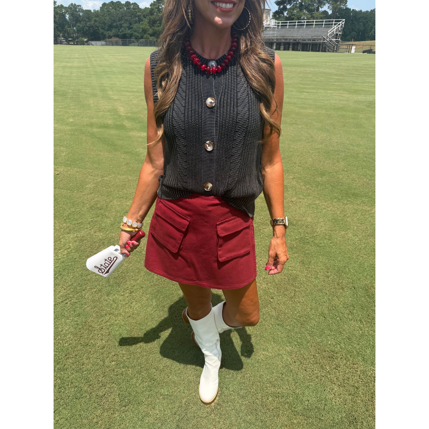A woman stands on a grassy golf course wearing a dark gray ribbed sleeveless button-front top, a burgundy mini skirt with front flap pockets, and white cowboy boots. She is holding a white golf club head cover with the brand name 'Ryder' visible.