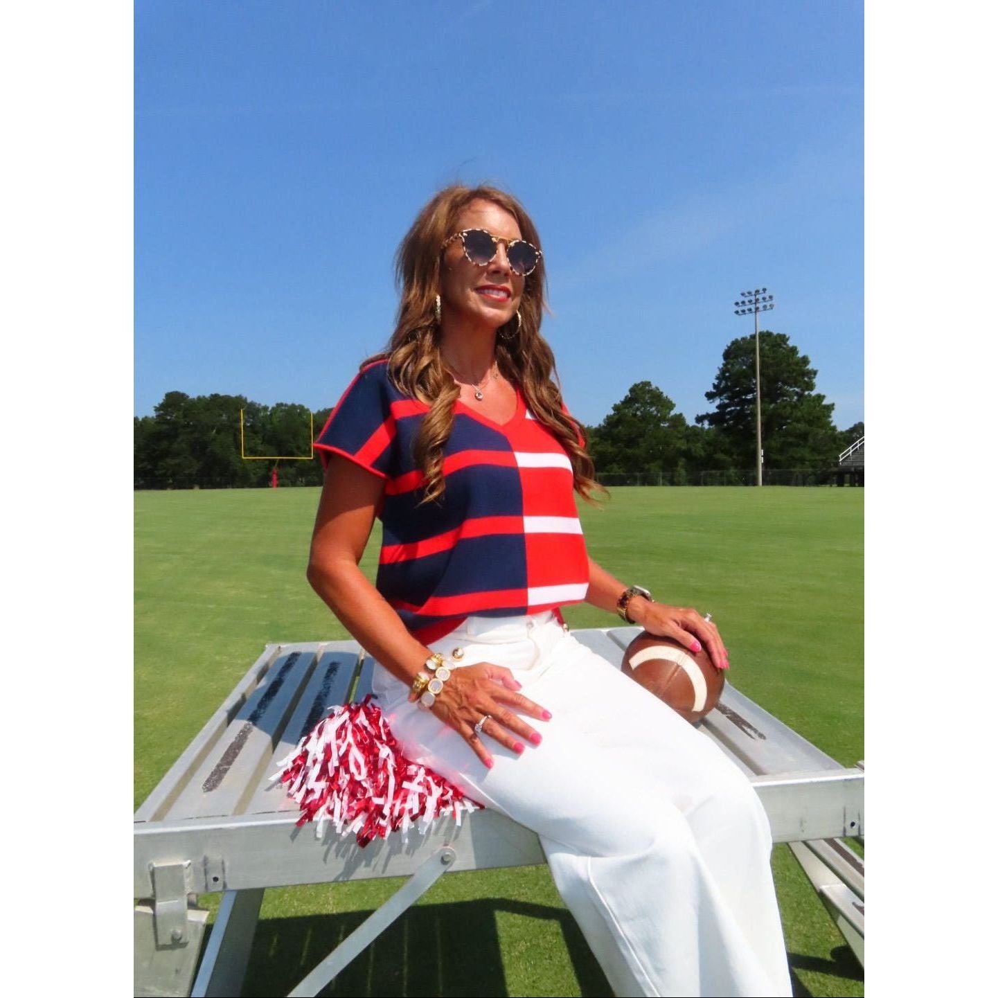 A woman wearing a red, navy, and white striped short-sleeve top and white pants sits on a metal picnic table on a grassy field, holding a football under a clear blue sky.