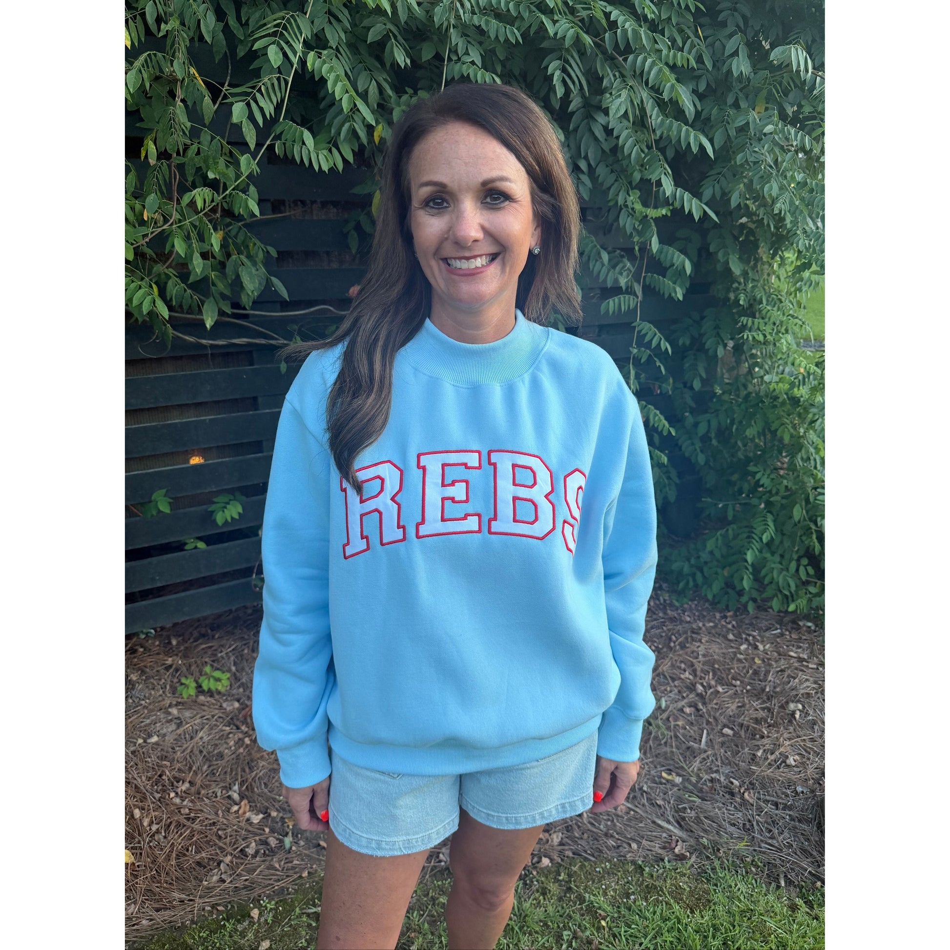 A woman standing outdoors in front of green foliage and a dark fence, wearing a light blue crewneck sweatshirt with 'REBS' printed across the chest in red and white, paired with light denim shorts.